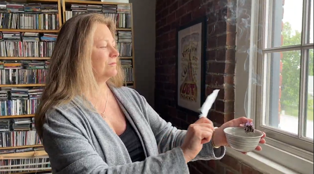 Load video: deb swingholm of flowering moon demonstrates space clearing by smudging with mountain sage and lavender with a white feather fan in a loft with brick walls